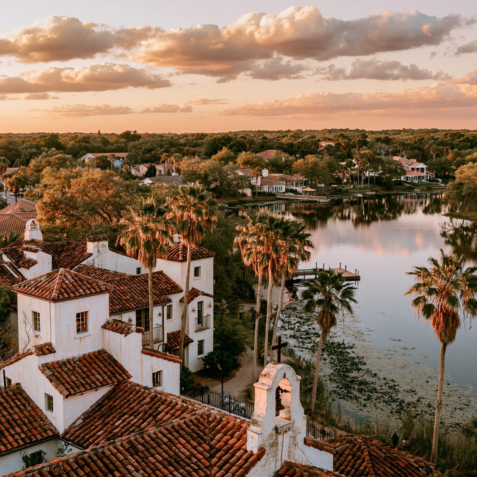 Golden-hour view of Avon Park, Florida - Mediterranean red-tile rooftops and a mission bell tower beside a calm palm-lined lake